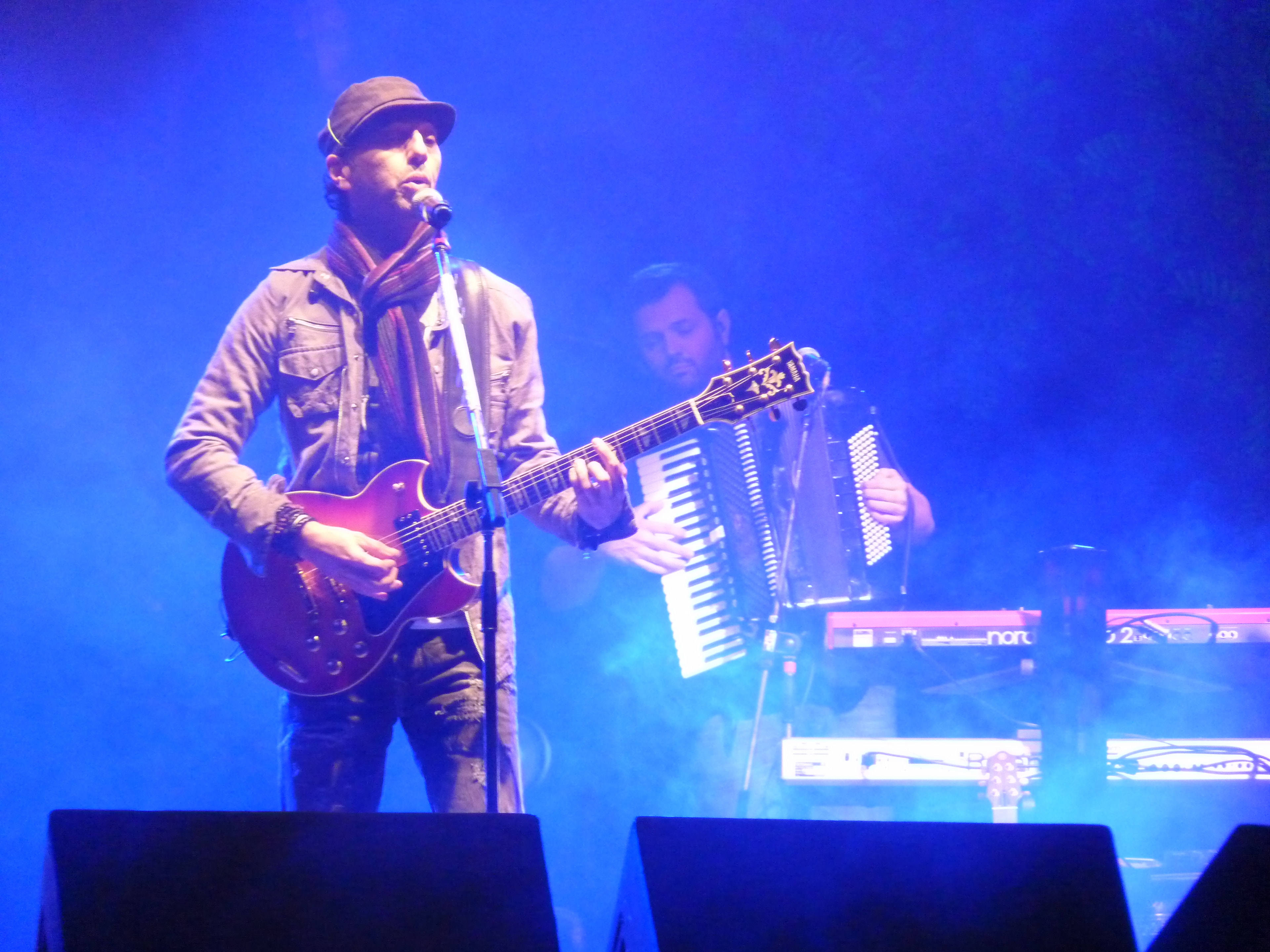 Músico brasileiro Zeca Baleiro sentado em uma cadeira de madeira, com violão na mão, sorriso tranquilo e fundo de ambiente urbano.