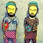 Dois homens jovens, Gustavo e Otavio Pandolfo, sentados em uma sala de estúdio, com papéis e lápis espalhados ao redor, sorriem para a câmera.