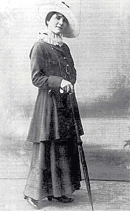 Mulher jovem com expressão intensa e cabelos cacheados, sentada em um ambiente literário com livros e papéis espalhados, inspirada em estilos Art Nouveau.