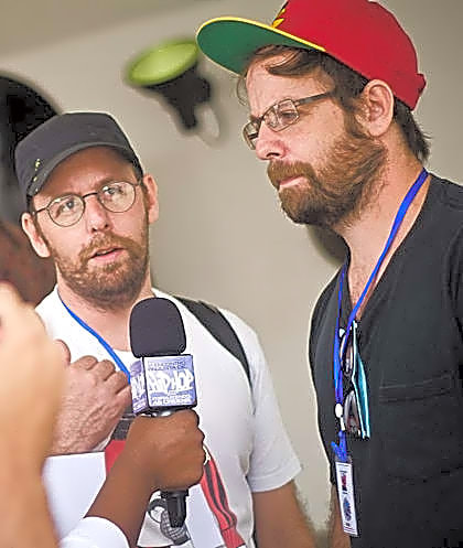 Dois homens idênticos, com barbas longas e vestidos de estilo clássico, sentados em uma paisagem natural, com fundo azul e nuvens brancas.