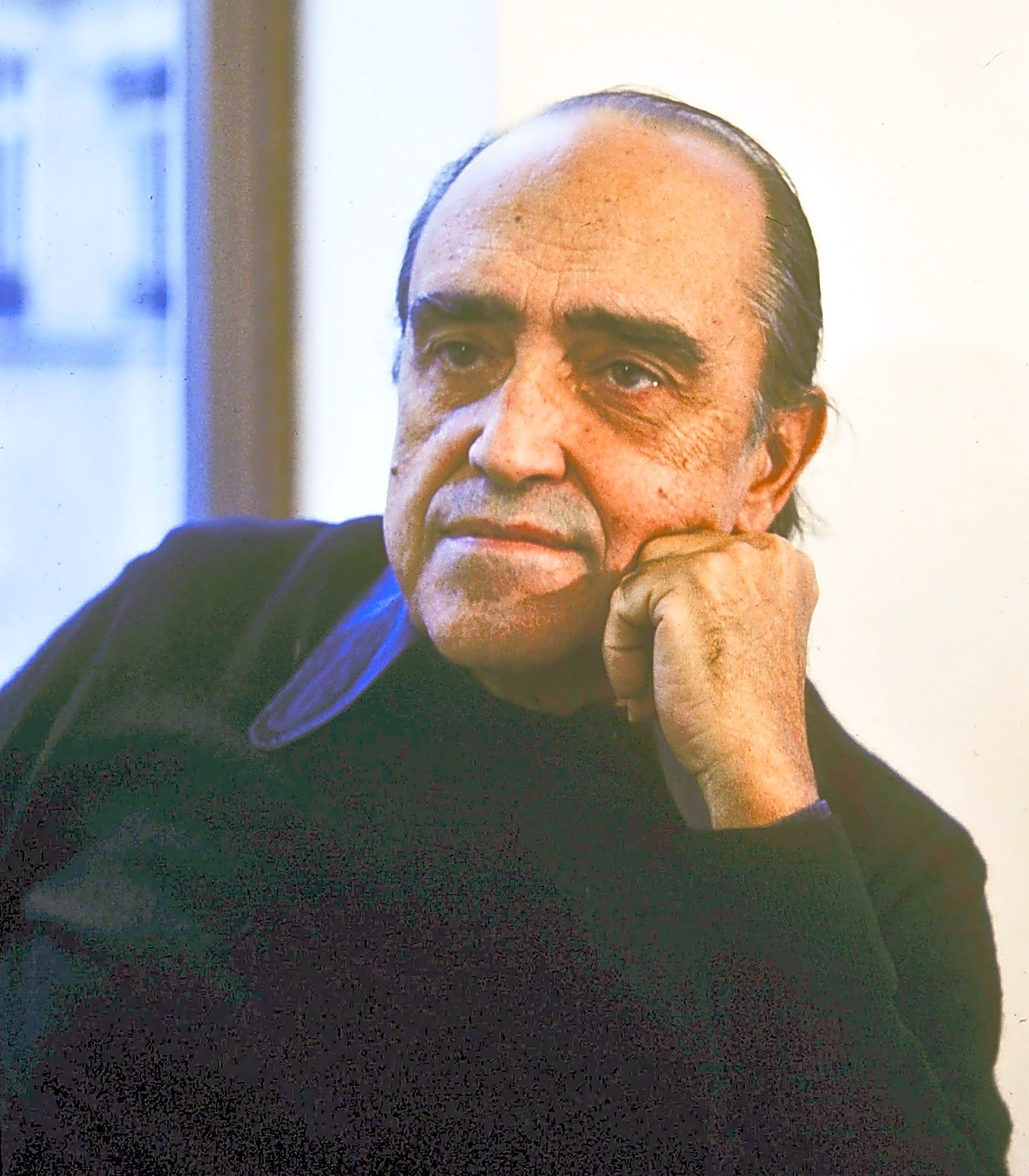 Arquiteto modernista brasileiro Oscar Niemeyer sentado em frente a um modelo de sua obra, com uma vista da cidade ao fundo.