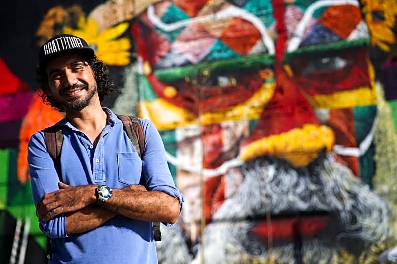 Pintura a óleo retratando Eduardo Kobra trabalhando em um mural urbano com cores vibrantes e detalhes realistas.