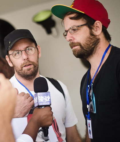 Dois artistas brasileiros, OSGEMEOS, sentados em uma escultura urbana de madeira, com expressões faciais misteriosas e cores vibrantes.